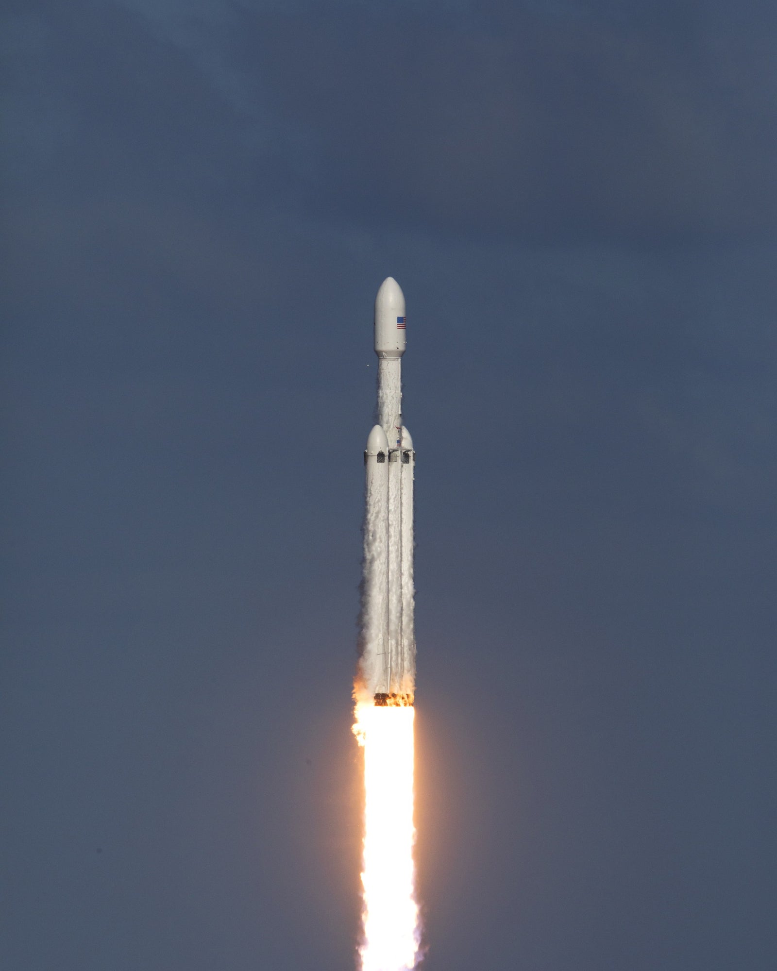 Falcon Heavy ascent. Triple cores and a white-hot plume against a dark sky.
